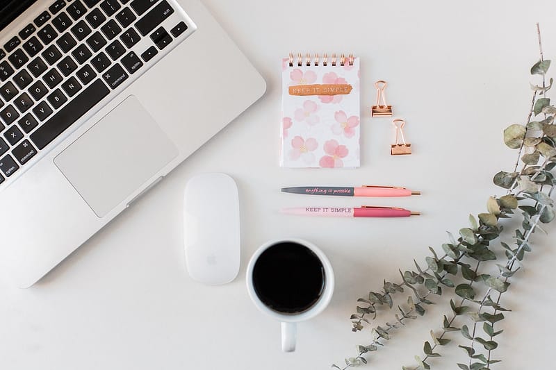 Rachel Jackson Marketing Tech Specialist. a minimalist picture of an office desk with a laptop on it. A layout of a desk set up with a lap top and a coffee mug with a white mouse, and pink clips and some foliage.