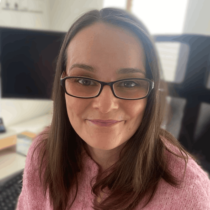 Headshot of Rachel Jackson - Marketing Tech Specialist, a woman with brown hair and glasses, wearing a pink jumper, smiling at the camera from her home office desk with natural light coming through the window behind her.
