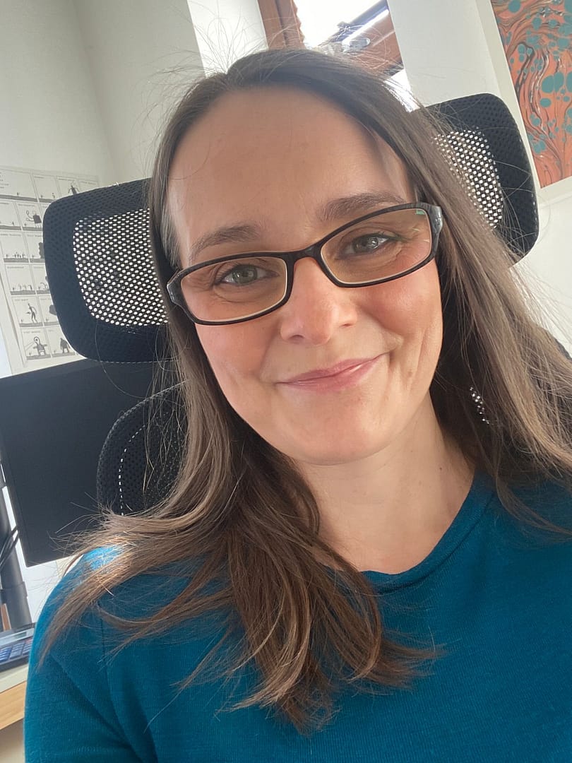 Rachel Jackson, UK-based marketing tech specialist, at her desk. Smiling, wearing with black glasses, wearing a teal coloured top.